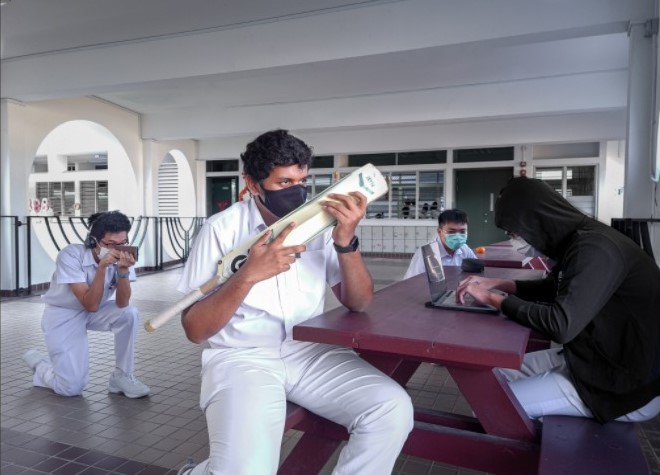 A masked student in white uniform holds a cricket bat while others sit or crouch nearby at a school corridor.